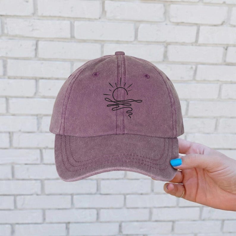 May include: A dusty rose baseball cap with a black embroidered design of a sun setting over water. The cap has a curved brim and a fabric strap closure. The hat is held by a person with blue nail polish.