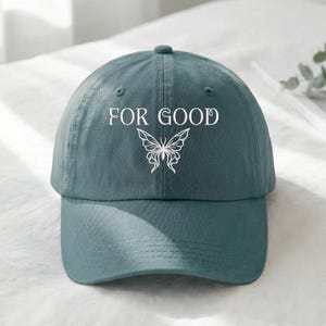 May include: A teal baseball cap with the words "FOR GOOD" and a butterfly graphic in white. The cap has a curved brim and a fabric strap closure on the back. The hat is made of a soft, durable material.