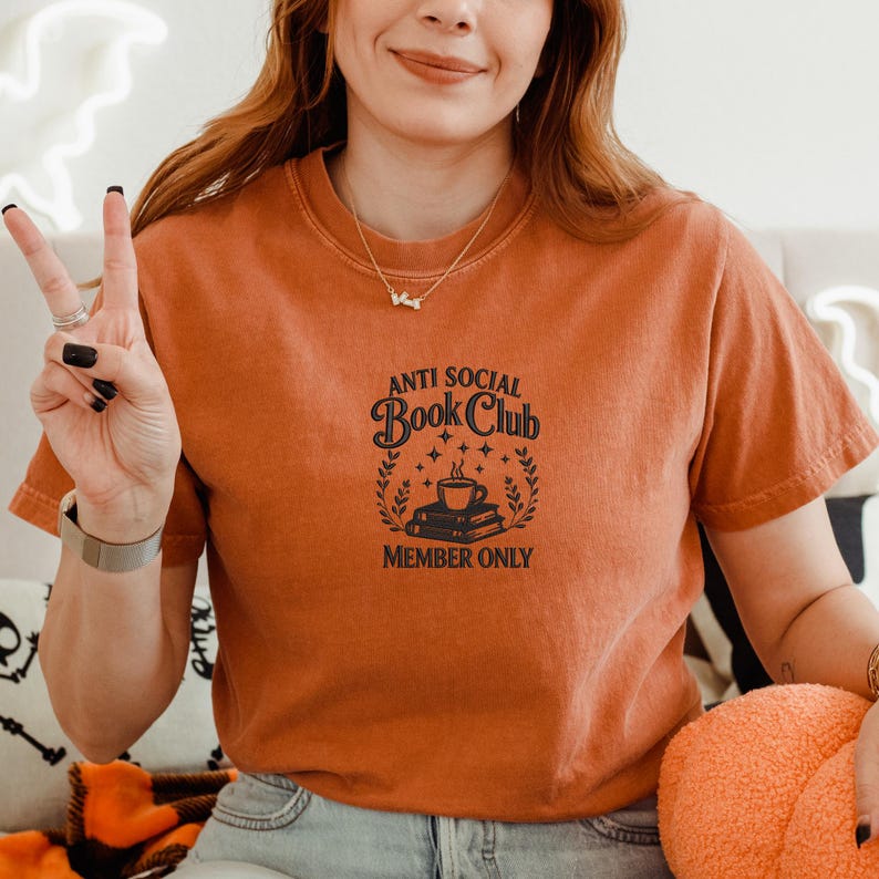 May include: Orange short-sleeved t-shirt with the text "ANTI SOCIAL Book Club MEMBER ONLY" in black. The graphic includes a coffee cup, books, stars, and a laurel wreath design. The shirt is a solid color.
