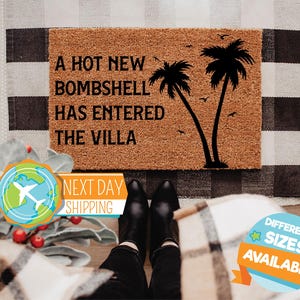 May include: A rectangular brown coir doormat featuring black text that reads A HOT NEW BOMBSHELL HAS ENTERED THE VILLA. The design includes a black silhouette of two palm trees and several small birds flying in the upper right corner of the mat.