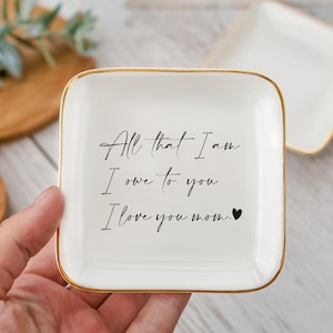 May include: A white square trinket dish with gold trim. The dish has the inscription "All that I am I owe to you I love you mom." in black script. A second dish is visible in the background.