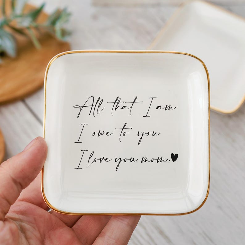 May include: A white square trinket dish with gold trim. The dish has the inscription "All that I am I owe to you I love you mom." in black script. A second dish is visible in the background.
