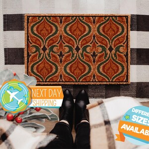 May include: A rectangular doormat with a repeating pattern of orange and green stylized floral shapes. The mat has a brown border and is placed on a black and white checkered rug. The words "NEXT DAY SHIPPING" are visible.