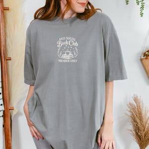 May include: Gray t-shirt with the text "ANTI SOCIAL Book Club MEMBER ONLY" in white. The design includes a stack of books, a coffee cup, and laurel leaves. The shirt has short sleeves and a crew neck.
