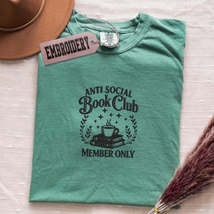 May include: A teal t-shirt with the text "ANTI SOCIAL Book Club MEMBER ONLY" in black. The design includes a coffee cup, books, and laurel leaves. A brown hat and a tag that says "EMBROIDERY" are in the background.