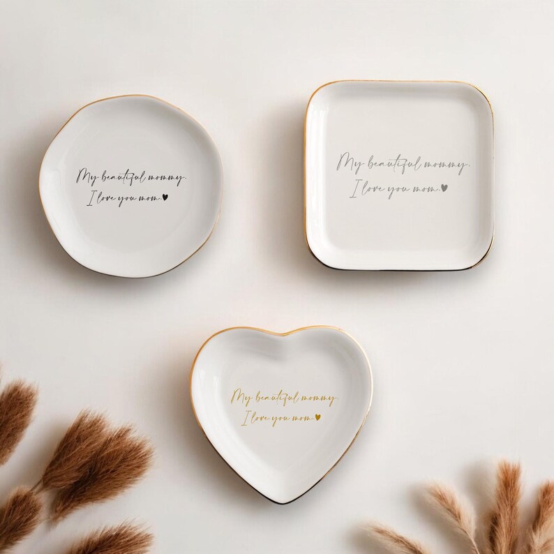 May include: Three white ceramic trinket dishes with gold rims. One is round, one square, and one heart-shaped. Each dish has the text "My beautiful mommy, I love you mom" in a handwritten style.