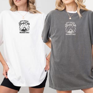 May include: Two oversized t-shirts, one white and one gray, featuring the text "ANTI SOCIAL Book Club MEMBER ONLY" in a circular design with books, a coffee cup, and stars. The shirts have short sleeves and a relaxed fit.