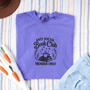 May include: A lavender t-shirt with the text "ANTI SOCIAL Book Club MEMBER ONLY" in black. The graphic includes a coffee cup, books, stars, and laurel leaves. The shirt is laid flat on a striped surface.