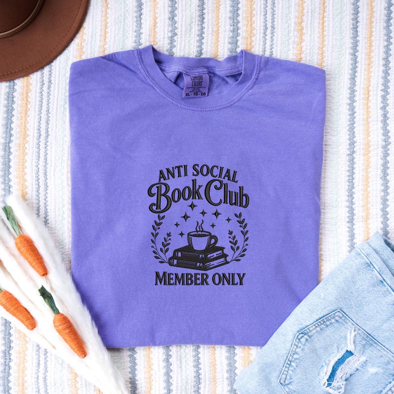 May include: A lavender t-shirt with the text "ANTI SOCIAL Book Club MEMBER ONLY" in black. The graphic includes a coffee cup, books, stars, and laurel leaves. The shirt is laid flat on a striped surface.