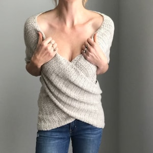 May include: A light gray knitted sweater with a draped, wrap-style design. The sweater has a deep V-neckline and a relaxed fit.