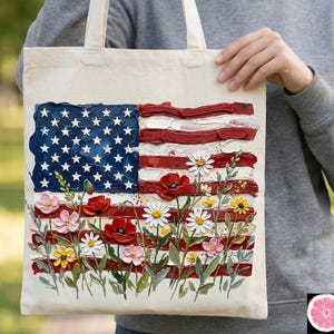 May include: Cream-colored tote bag featuring a textured painting of the American flag with a floral border. The flag design includes blue with white stars, and red and white stripes. The floral border includes red poppies, white daisies, and other colorful flowers.