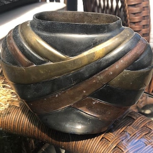 May include: A black, gold and copper coloured ceramic vase with a woven design. The vase is sitting on a wicker surface.