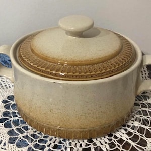 Peut inclure: Un plat à casserole en céramique beige avec un couvercle. Le plat a deux poignées et une surface tachetée. Le couvercle présente une bande décorative marron et un petit bouton rond. Le plat à casserole est conçu pour la cuisine et le service.