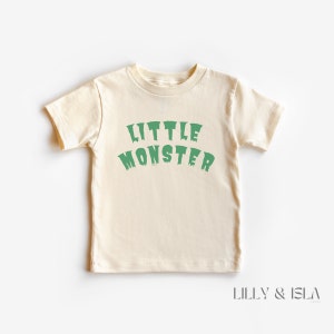 May include: A cream coloured short-sleeved t-shirt with the text "LITTLE MONSTER" in green dripping letters.