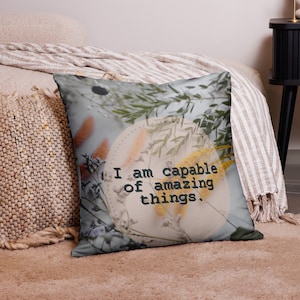 May include: Square decorative pillow with a floral design and the text "I am capable of amazing things." The pillow features a light blue background with dried flowers in shades of yellow, orange, and green.