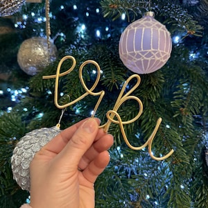 May include: A gold wire word spelling "love" is held in front of a Christmas tree. The tree is decorated with silver and purple baubles and white lights. The word art is in a cursive font.