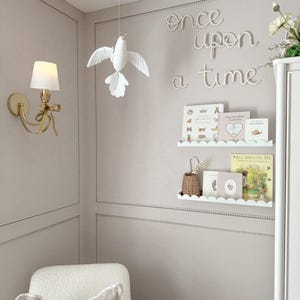 May include: A nursery interior featuring a white dove mobile, a gold wall sconce, and a white armchair. The wall displays the phrase "once upon a time". Two white shelves hold books and a small basket.