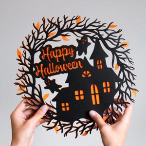 Könnte beinhalten: Schwarze Halloween-Dekoration mit einer Silhouette eines Spukhauses und orangefarbenen Akzenten. Das Design umfasst den Text "Happy Halloween", Geister und einen Baumzweig-Rand. Die Dekoration wird von zwei Händen gehalten.