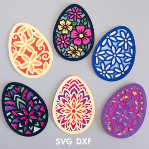 Op de afbeelding: Zes verschillende paasei-ontwerpen met ingewikkelde uitsnijdingen. De eieren zijn in verschillende kleuren, waaronder geel, oranje, blauw, paars, roze en groen. De ontwerpen omvatten bloemmotieven, geometrische vormen en spiralen. De tekst "SVG DXF" staat onderaan de afbeelding.