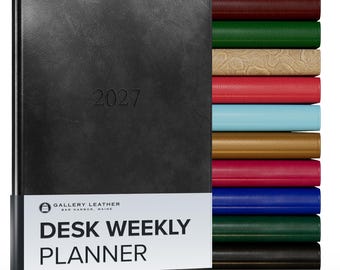 May include: A black 2027 desk weekly planner with a white paper band featuring the text Gallery Leather Bar Harbor, Maine Desk Weekly Planner. To the right, a vertical stack of planners in various colors including burgundy, green, tan, red, blue, gold, pink, navy, dark green, and black.