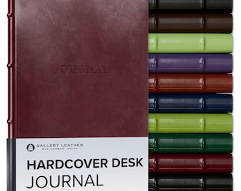 May include: A burgundy hardcover desk journal with the word JOURNAL embossed on the front cover. A white paper band features the text GALLERY LEATHER BAR HARBOR, MAINE HARDCOVER DESK JOURNAL. A stack of journals in various colors is visible behind.