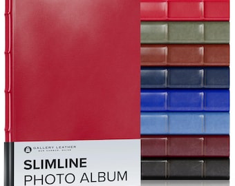 May include: A red Slimline Photo Album by Gallery Leather Bar Harbor Maine stands upright, featuring a smooth cover and a spine with raised bands. To its right, a stack of eight additional albums in red, green, brown, navy, blue, light blue, burgundy, and black.