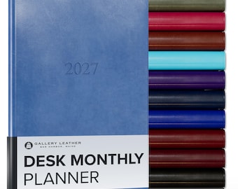 May include: A blue 2027 desk monthly planner with a white paper band featuring the text Gallery Leather Bar Harbor, Maine Desk Monthly Planner. A stack of additional planners in various colors including olive, red, brown, teal, and black is visible.
