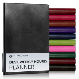 May include: A black 2027 desk weekly hourly planner with a white paper band featuring the text Gallery Leather Bar Harbor, Maine Desk Weekly Hourly Planner. A stack of additional planners in various colors including red, pink, brown, purple, blue, green, and black is visible to the right.
