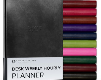 May include: A black 2027 desk weekly hourly planner with a white paper band featuring the text Gallery Leather Bar Harbor, Maine Desk Weekly Hourly Planner. A stack of additional planners in various colors including red, pink, brown, purple, blue, green, and black is visible to the right.
