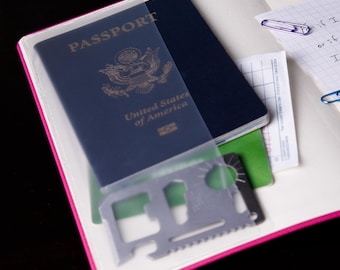 May include: A dark blue United States passport inside a clear plastic sleeve of a pink notebook. A silver metal multi-tool card with various cutouts and a serrated edge rests on top. A piece of grid paper with purple and blue paperclips is visible.