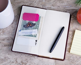 May include: An open notebook with a dark cover containing a US dollar bill, a pink ticket, a moth sticker, a fortune cookie slip reading All that we are is a result of what we have thought, an ace of spades playing card, and a black fountain pen.