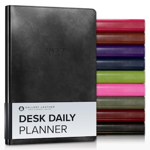 May include: A black 2027 desk daily planner with a white paper band featuring the text GALLERY LEATHER BAR HARBOR, MAINE DESK DAILY PLANNER. A stack of additional planners in various colors including red, brown, purple, blue, green, and pink is visible.