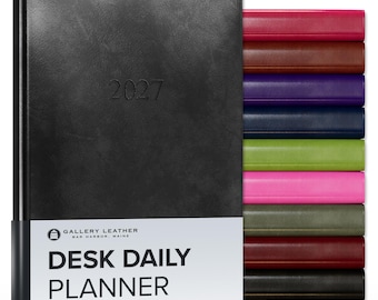 May include: A black 2027 desk daily planner with a white paper band featuring the text GALLERY LEATHER BAR HARBOR, MAINE DESK DAILY PLANNER. A stack of additional planners in various colors including red, brown, purple, blue, green, and pink is visible.