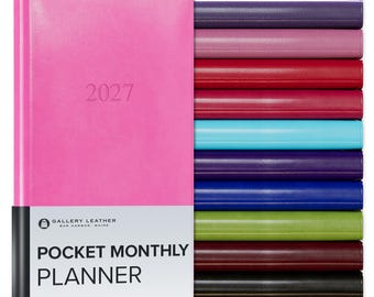 May include: A bright pink 2027 pocket monthly planner by Gallery Leather with a white paper band featuring the text Gallery Leather Bar Harbor Maine Pocket Monthly Planner, stacked next to a vertical row of planners in various solid colors.
