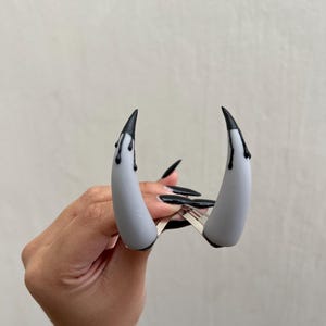 Op de afbeelding: Een paar grijze, hoornvormige haarclips met zwarte punten en druipende accenten. De clips worden op hun plaats gehouden door een zilveren metalen sluiting. Close-up van de haaraccessoires.