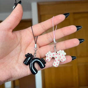 Puede incluir: Dos colgantes hechos a mano. Uno es negro y con forma de caballo, el otro es blanco con corazones rojos. Ambos tienen cierres metálicos plateados y cordones negros o rosas. Los colgantes se sostienen sobre un fondo neutro.
