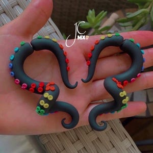 Puede incluir: Un par de pendientes negros en forma de tentáculos curvos con ventosas de colores arcoíris. Los pendientes están hechos de un material liso y se muestran sobre un fondo neutro. Los pendientes tienen un diseño artístico único.