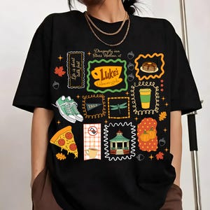 May include: A black short-sleeve t-shirt featuring a collage of colorful graphics including a Luke's Diner logo, a stack of pancakes, a coffee cup, a slice of pepperoni pizza, a gazebo, pumpkins, green sneakers, and the text Dragonfly Inn Stars Hollow.