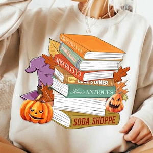 May include: A beige crewneck sweatshirt featuring a graphic of a stack of books with titles Dragonfly Inn, Doose's Market, Miss Patty's, Luke's Diner, Kim's Antiques, and Soda Shoppe, accented by a purple witch hat and two orange jack-o'-lanterns.
