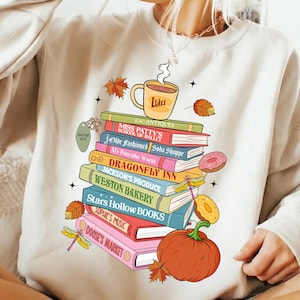 May include: A beige crewneck sweatshirt featuring a graphic of a stack of colourful books topped with a coffee mug labelled Luke's. The book spines display text including Miss Patty's School of Ballet, Dragonfly Inn, and Doose's Market, with autumn leaves, a pumpkin, and dragonflies.