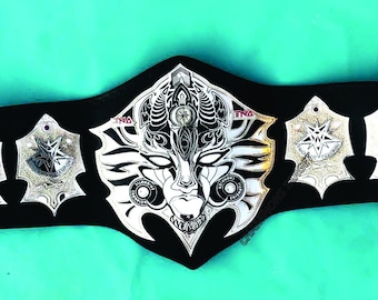 May include: A black leather belt with silver metal accents. The belt has a large, ornate, silver metal buckle with a detailed design. The buckle features a face with a crown and a geometric pattern.