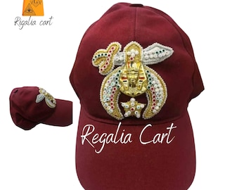 Puede incluir: Una gorra de béisbol de color granate con un diseño bordado en oro, blanco y verde que representa una luna creciente, una estrella y una espada. La gorra tiene el texto "Regalia Cart" bordado en la parte delantera.