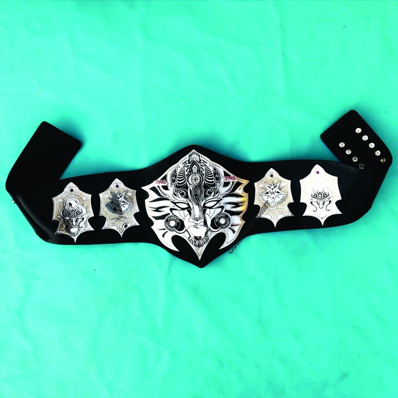 Puede incluir: Un cintur&oacute;n de cuero negro con detalles en metal plateado. El cintur&oacute;n tiene una hebilla grande, ornamentada, de metal plateado con un dise&ntilde;o detallado. La hebilla presenta una cara con una corona y un patr&oacute;n geom&eacute;trico.