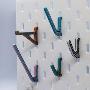 Può includere: Cinque ganci in plastica colorati a forma di V, montati su una parete di bacheca bianca. I ganci sono in tonalità arancio, rosa, blu, turchese e nero.