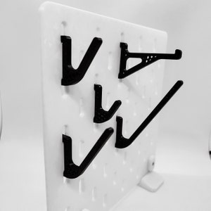 Può includere: Un pannello perforato bianco con quattro ganci in plastica nera. I ganci sono di forme e dimensioni diverse.