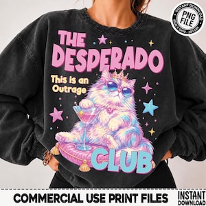 May include: A charcoal gray crewneck sweatshirt featuring a graphic of a fluffy cat wearing sunglasses and a crown, sitting on a pink cushion while holding a martini glass. The text reads THE DESPERADO, This is an Outrage, and CLUB.