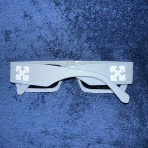 Puede incluir: Gafas de sol blancas con montura rectangular y lentes negras. Las patillas presentan un logotipo decorativo. Las gafas de sol están sobre un fondo azul.