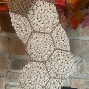 May include: A handmade Christmas stocking with a crochet design. The stocking features a brown ribbed cuff and a body composed of beige hexagonal crochet flowers. The flowers are outlined with brown yarn. The stocking is hanging in front of a brick fireplace.