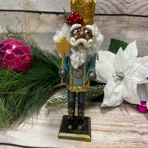 May include: A decorative Christmas nutcracker figurine. The nutcracker is wearing a gold glitter crown, a teal jacket with gold trim, and a white beard. It holds a gold star wand and stands on a square base. Red berries adorn the crown.