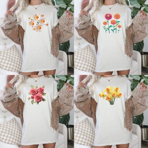 Op de afbeelding: Vier witte shirts met lange mouwen met verschillende bloemenprints. De shirts hebben een boeket witte madeliefjes, een bos rode rozen, een bos gele narcissen en een groep oranje en gele bloemen.